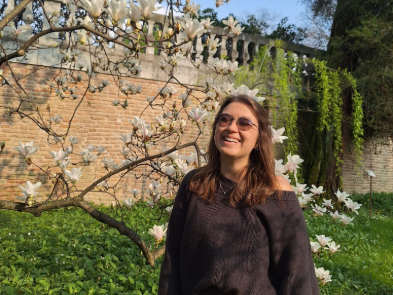 Eine Frau mit längeren Braunen Haaren und getönter Brille lacht fröhlich. Im Hintergrund blüht ein Baum vor einer Ziegelmauer.