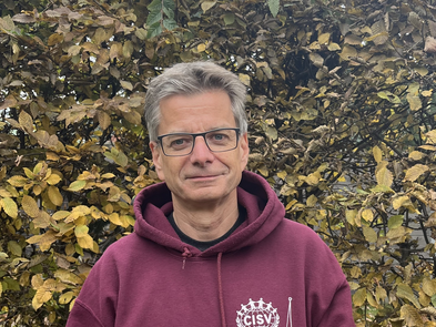 Mann mit leicht grauen Haaren und Brille steht vor einer BlÀtterwand. Er trÀgt einen weinroten Kapuzenpullover.