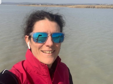 Selfie einer Frau an einem See. Sie trägt eine Sonnenbrille, ein rotes Gilet und lächelt in die Kamera.