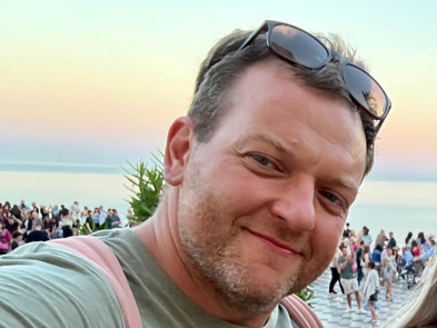 Selfie von einem lĂ€chelnden Mann vor einem Strand voller Menschen. Er trĂ€gt eine Sonnenbrille nach oben geschoben, einen Rucksack und einen grĂŒnes T-Shirt.