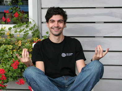 Junger Mann sitzt im Schneidersitz vor einer Hauswand. Links von ihm sind rote Blumen zu sehen. Er trägt eine blaue Jeans und ein schwarzes T-Shirt