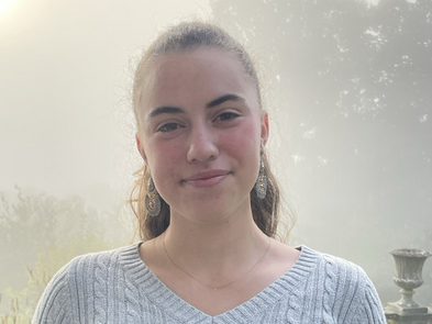 Junge Frau lächelt in direkt in die Kamera. Sie trägt einen hellgrauen Strickpullover und die braunen Haare in einem Pferdeschwanz zusammengebunden.