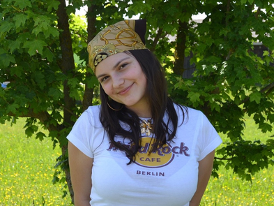 Junge Frau hat ihre Arme hinter dem Rücken verschränkt und lächelt mit leicht geneigten Kopf in die Kamera. Sie trägt ein gelbes Tuch am Kopf und ein weißes Hard Rock Café T-Shirt.