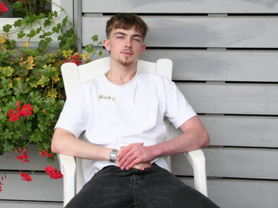 Junger Mann sitzt lässig auf einem weißen Plastikstuhl vor einer Hauswand. Links von ihm sind rote Blumen zu sehen. Er trägt ein weißes T-Shirt und eine schwarze Hose.