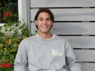 Junger Mann sitzt vor einer Hauswand und lächelt in die Kamera. Links neben ihm sind rote Blumen zu sehen. Er trägt einen hellgrauen CISV-Pullover.